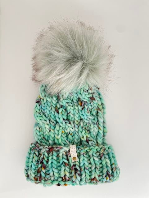 Teal knit hat with colorful pattern and gray fur pom-pom on a white background